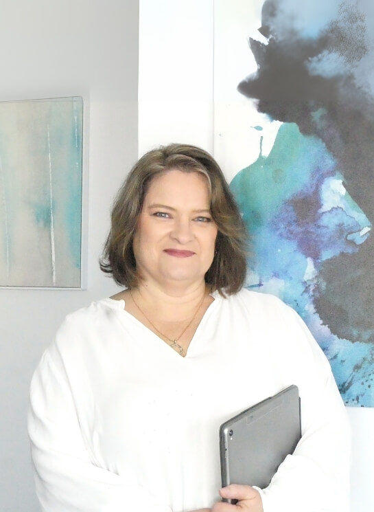 A professional woman wearing a white blouse is smiling while holding a tablet, standing in front of abstract blue artwork in a modern office.