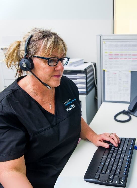 Female healthcare receptionist wearing a headset and glasses, working at a computer in an office setting.